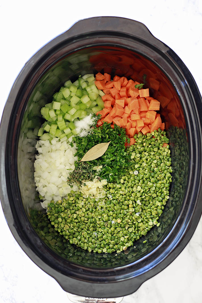 A crock pot filled with the ingredients for split pea soup, including carrots, celery, herbs, onions and dried split peas. 