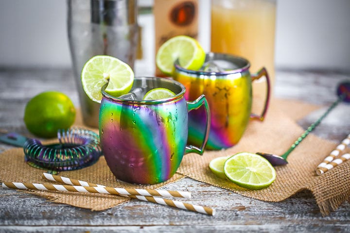 Two Moscow Mules served in rainbow mugs with lime slices with a cocktail shaker, striped straws, ginger beer, and vodka scattered around.