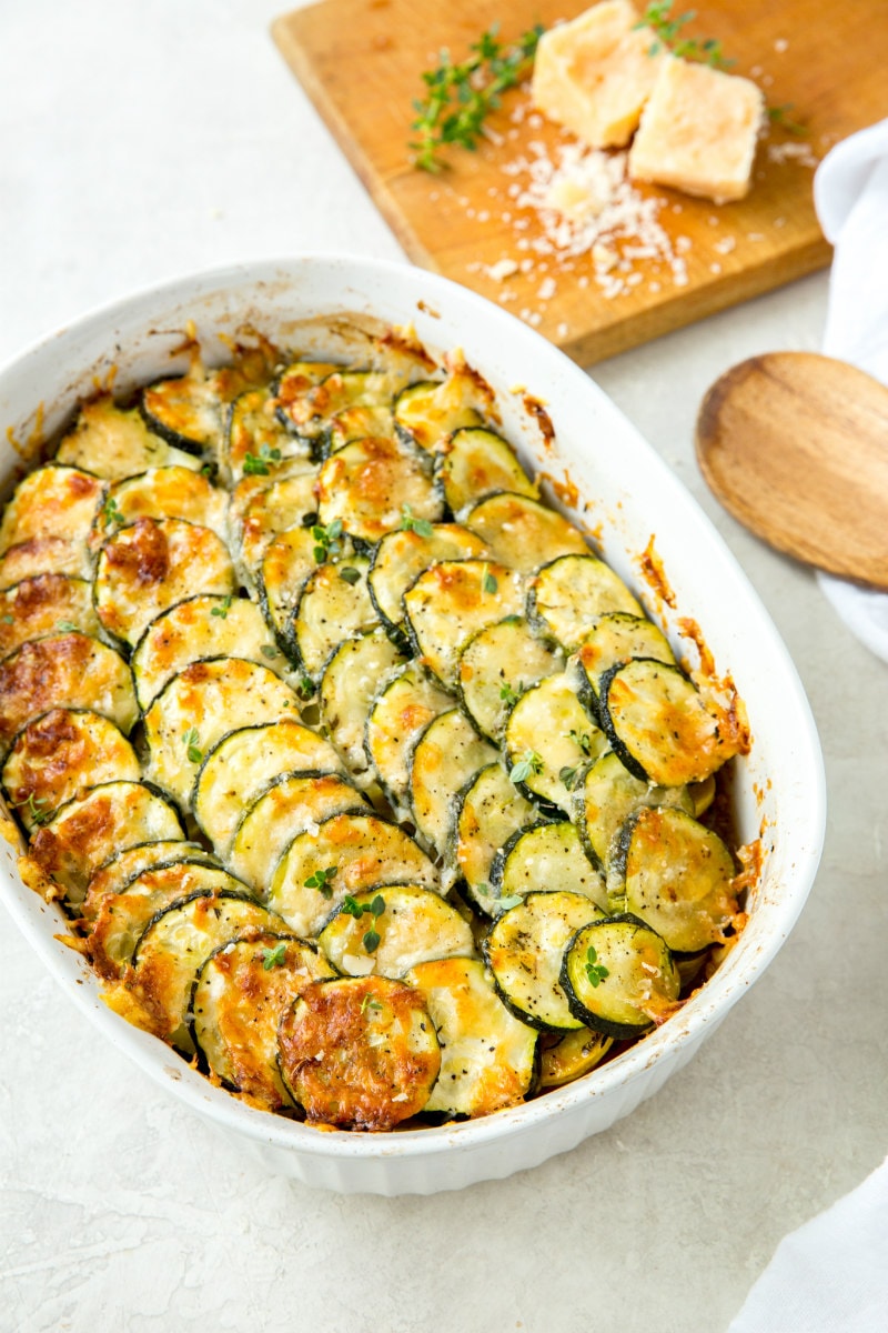 zucchini and tomato gratin in a white casserole dish with a wooden cutting board with cheese on it and a wooden spoon on the side